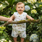 Smiling baby wearing a white multi printed muslin jabla shirt and shorts set, standing outdoors in a garden.