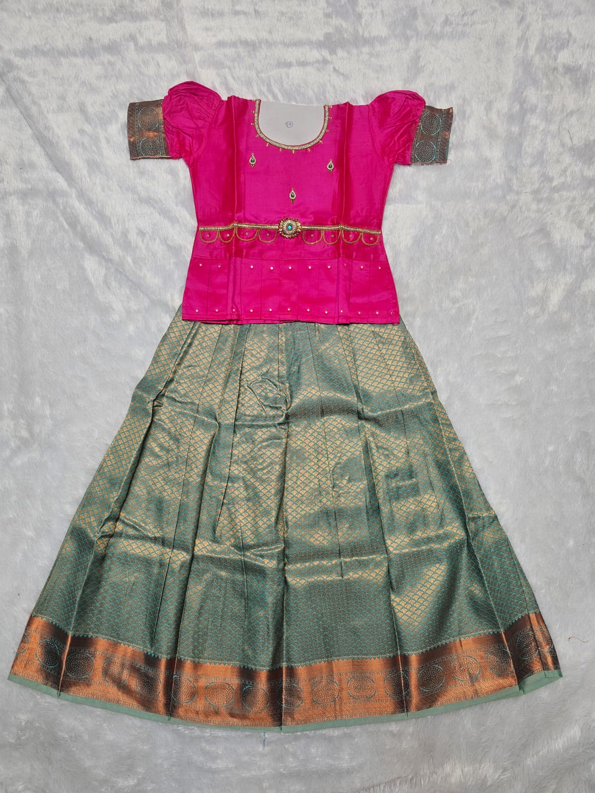 Bright pink semi silk blouse with teal green bridal silk skirt pattu pavadai for girls featuring puff sleeves, waist belt design and double chain neck stone work